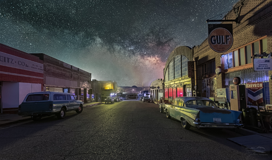 Milky Way and stars shine bright over a small town street lined with historic cars.